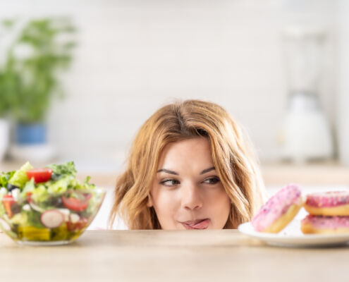 Beautiful young woman tempted having to make choice between fresh lettuce salad and doughnut in healthy unhealthy food, detox eating, calories and diet concept.