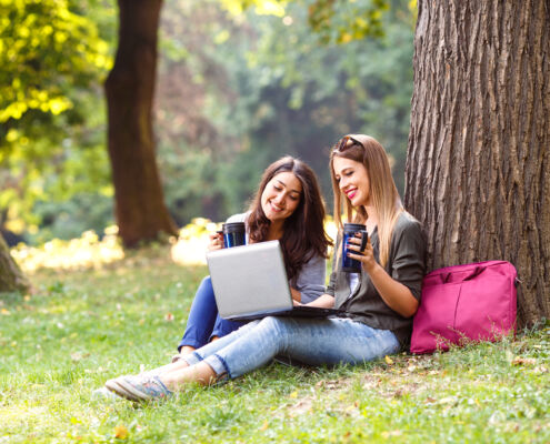 Portrait of two female student sitting in park with laptop.