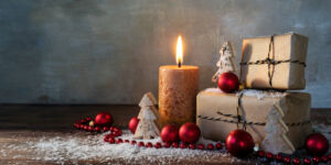 Geschenke Advent Weihnachten; two gift boxes and a burning candle decorated with red christmas baubles and small wooden toy trees in some snow on rustic wood, vintage background with large copy space, panorama format, selected focus, narrow depth of field